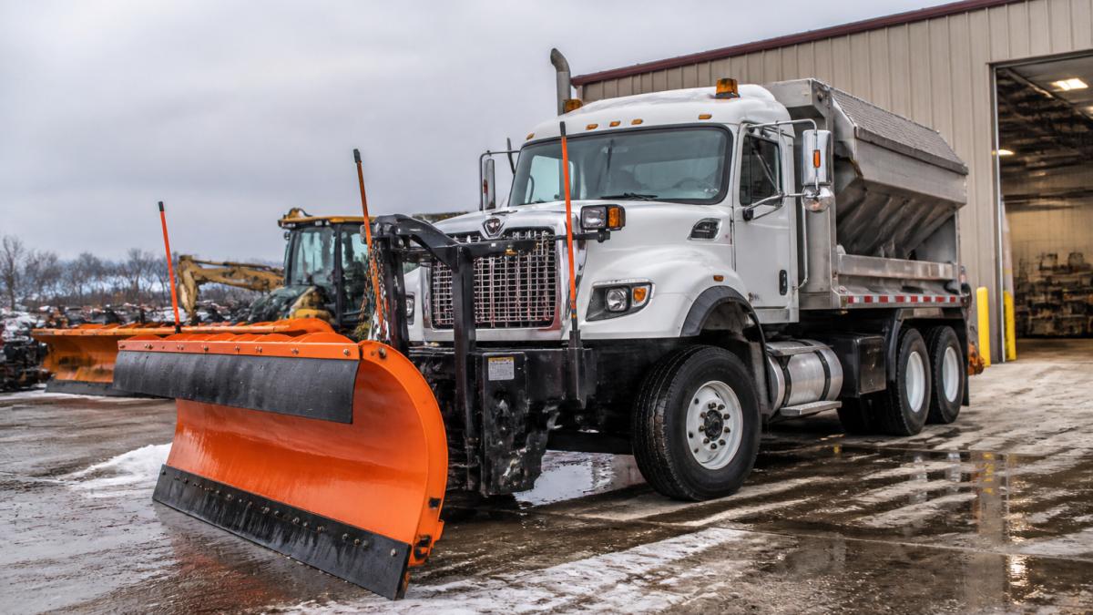 Professional snowplow truck with modern plow blade and salt spreader system attached, ready for winter operations in a service yard with clear equipment maintenance area